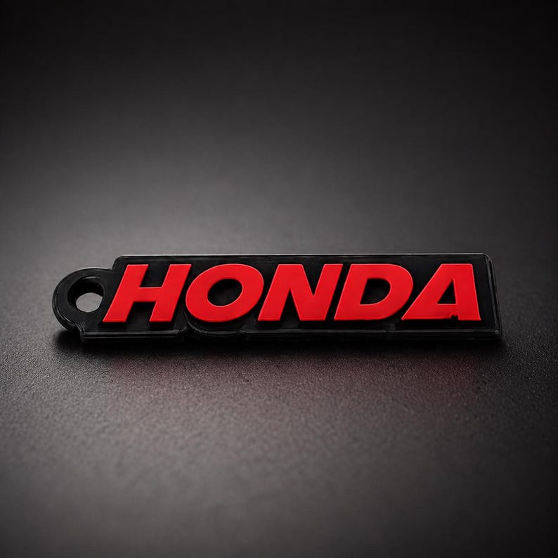 May include: A black and red Honda keychain. The word "HONDA" is in red, bold letters on a black rectangular background. The keychain has a small hole on the left side for attachment.