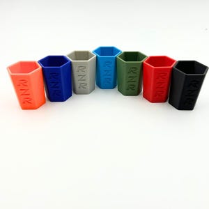 May include: A collection of seven hexagonal shot glasses in various colors: orange, blue, gray, light blue, green, red, and black. Each glass has the letters "RNR" printed on the side. The glasses are arranged in a row against a white background.