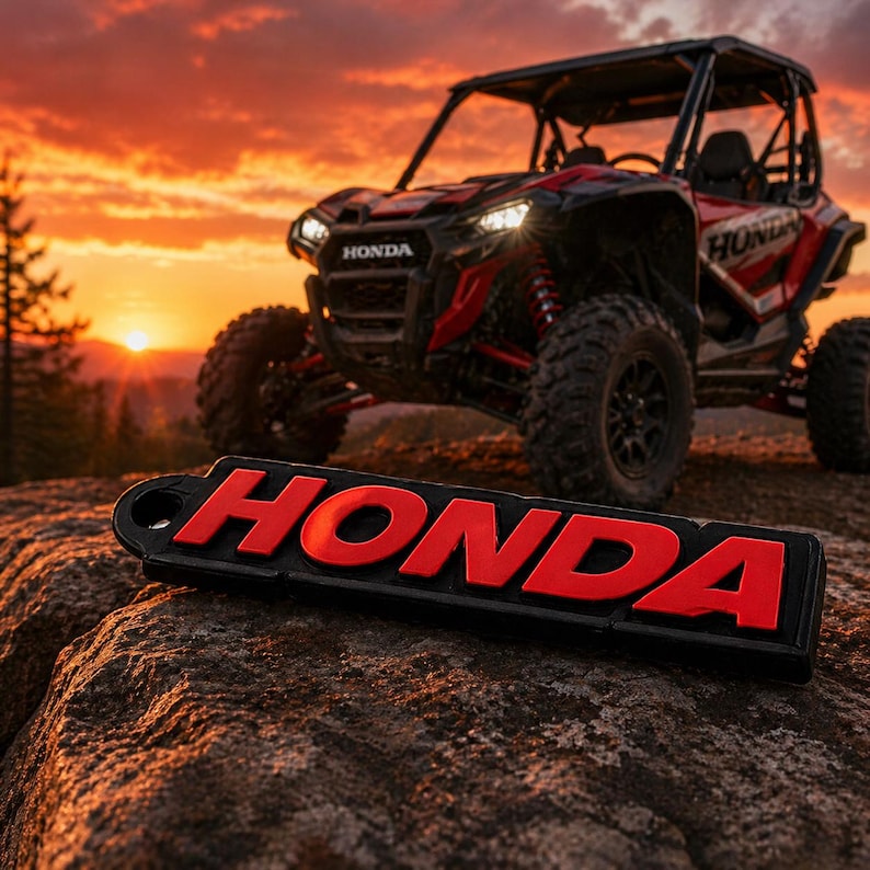 May include: A black and red Honda keychain with the word "HONDA" in red, resting on a rock. In the background, a red and black Honda UTV is parked, with a sunset in the background.