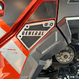 May include: A close-up of a silver metal plate with the words "GENERAL" and "G-O" on it. The plate is attached to a red and black vehicle.