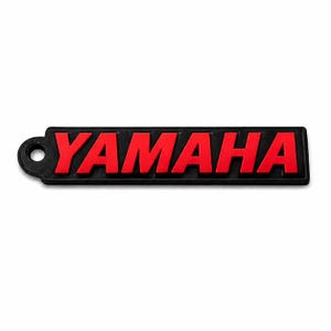 Pu&ograve; includere: Portachiavi rettangolare nero con la scritta "YAMAHA" in rosso in grassetto, carattere sans-serif. Il portachiavi ha un piccolo foro circolare sul lato sinistro. Lo sfondo &egrave; bianco.