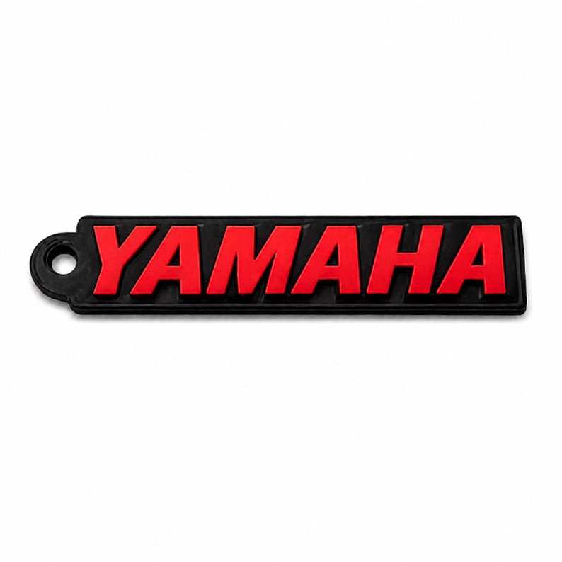 Pu&ograve; includere: Portachiavi rettangolare nero con la scritta "YAMAHA" in rosso in grassetto, carattere sans-serif. Il portachiavi ha un piccolo foro circolare sul lato sinistro. Lo sfondo &egrave; bianco.