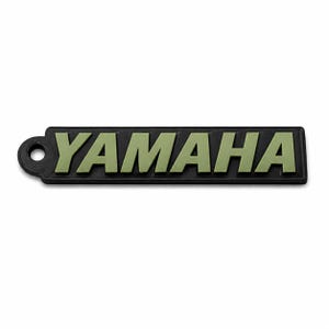 Pu&ograve; includere: Un portachiavi rettangolare nero con la scritta "YAMAHA" in lettere verde oliva. Il portachiavi ha un piccolo foro circolare sul lato sinistro.