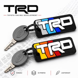 May include: Two rectangular keychains featuring the TRD logo in white lettering on a black background. One keychain has blue and grey accents, while the other has yellow, orange, and red accents. Each is attached to a grey key with the number 122.