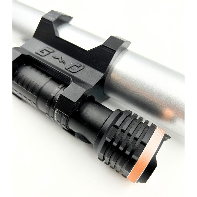 Może przedstawiać: Czarna latarka z żebrowaną głowicą i pomarańczowym akcentem, zamontowana na srebrnym cylindrycznym obiekcie. Uchwyt jest czarny i zawiera litery "G" i "P". Latarka jest przeznaczona do użytku taktycznego lub zewnętrznego.