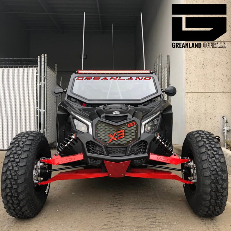 May include: A black off-road vehicle with red accents and a black grille with the text "X3" in red. The vehicle has a black roll cage and a black windshield with the text "GREANLAND" in red. The vehicle is sitting on a black asphalt surface.