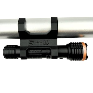 Może przedstawiać: Czarna latarka zamontowana na srebrnym słupie z czarnym wspornikiem. Latarka ma teksturowany uchwyt i miedziany pierścień w pobliżu soczewki. Wspornik ma litery "G", g&oacute;rę i kwadratowy symbol.