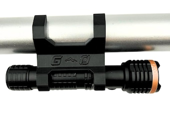 May include: Black flashlight mounted on a silver pole with a black bracket. The flashlight has a textured grip and a copper-colored ring near the lens. The bracket has the letters "G", a mountain, and a square symbol.
