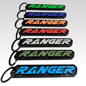 May include: A collection of keychains with the word "RANGER" in various colors: green, blue, orange, red, white, olive green, and light blue. Each keychain has a black border and a black keyring.