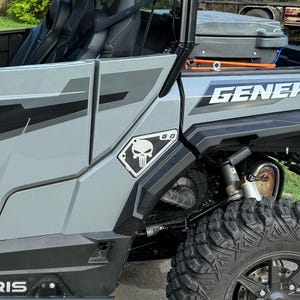 May include: A gray off-road vehicle with a black skull and crossbones decal on the side. The decal is in a diamond shape and says "G.D." above the skull. The vehicle has a black roll cage and a black and gray interior.