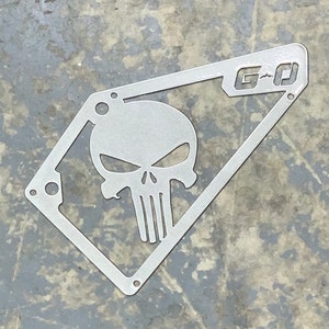May include: A silver metal cutout of a skull with the text "G-D" in the upper right corner. The skull is a stylized version of the Punisher logo.