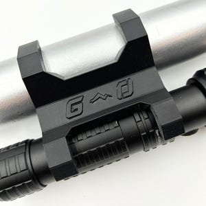 Może przedstawiać: Czarny metalowy uchwyt z literami "G" i stylizowanym symbolem wygrawerowanym na nim, przymocowany do srebrnego cylindrycznego obiektu. Czarny cylindryczny obiekt jest przymocowany do uchwytu.