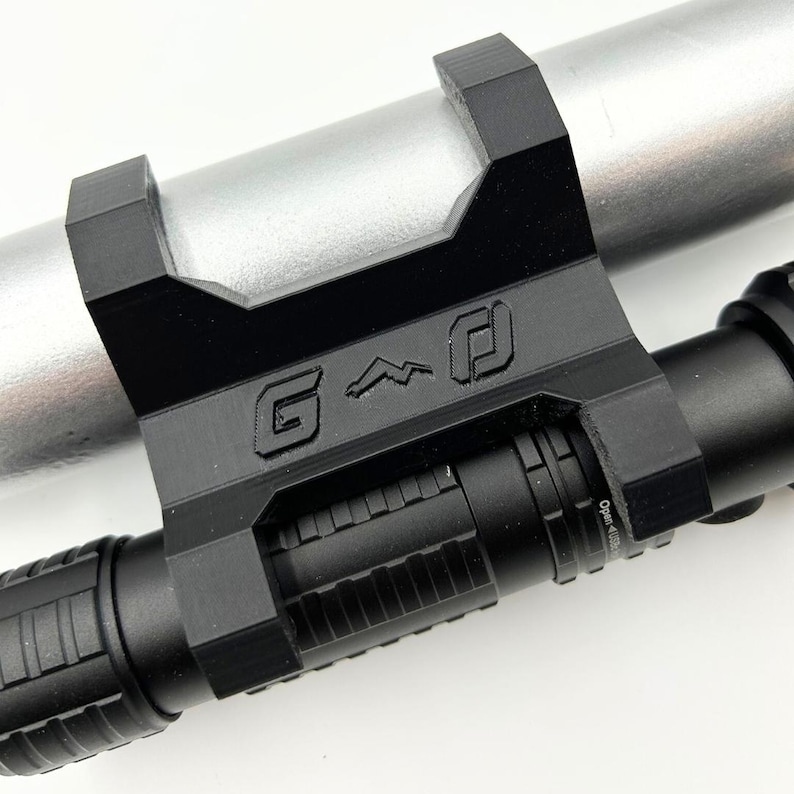 Może przedstawiać: Czarny metalowy uchwyt z literami "G" i stylizowanym symbolem wygrawerowanym na nim, przymocowany do srebrnego cylindrycznego obiektu. Czarny cylindryczny obiekt jest przymocowany do uchwytu.