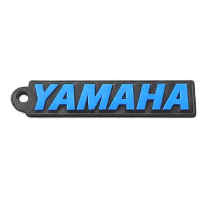 Pu&ograve; includere: Un logo Yamaha nero e blu. La parola "YAMAHA" &egrave; in lettere blu in rilievo su uno sfondo rettangolare nero. Il logo ha un piccolo foro sul lato sinistro.