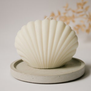 May include: A white seashell-shaped candle on a grey concrete base. The candle is made to look like a seashell with ridges and a smooth surface.