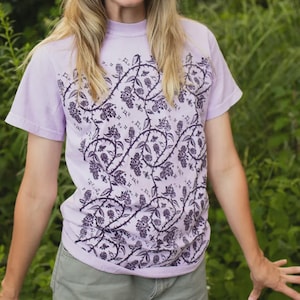 Op de afbeelding: Een lichtpaars t-shirt met korte mouwen en een donkerpaars herhalend patroon van doornige ranken, bladeren en kleine bloemen. Het shirt wordt gecombineerd met een olijfgroene korte broek en een bijpassende olijfgroene bucket hat met een ronde patch.