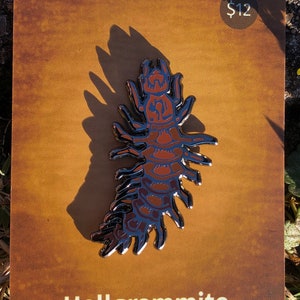 May include: A silver enamel pin shaped like a centipede with red and black details. The pin is on a brown card with the text "Hellgrammite Enamel Pin" and a price tag of $12.