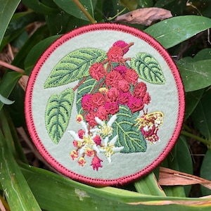 May include: A circular embroidered patch featuring a light sage green background with a detailed botanical design of red berries, green leaves, white flowers, and a small yellow butterfly, all enclosed within a dark red stitched border.