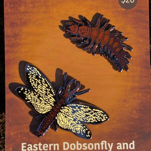 May include: A set of two enamel pins featuring a brown and black hellgrammite and a yellow and black eastern dobsonfly. The pins are on a brown card with the text "Eastern Dobsonfly and Hellgrammite Pin Set" and "$20".