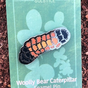 May include: Wooly Bear Caterpillar Enamel Pin by Solstice Handmade. The pin features a black, orange and white caterpillar against a black background. The pin is on a green card with the text "Solstice" and "Wooly Bear Caterpillar Enamel Pin" and the website "solsticehandmade.com".