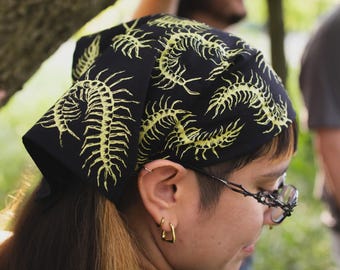 May include: A black bandana featuring a repeating pattern of yellow centipedes. The fabric is tied around the head, displaying the detailed insect illustrations across the surface. The design consists of multiple centipedes in various orientations.