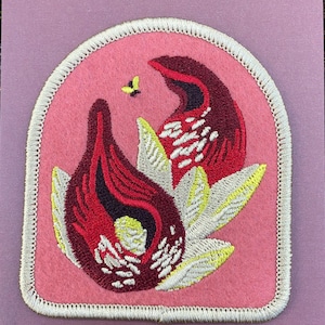 Könnte beinhalten: Ein rosa Bügeleisen-Patch mit einer gestickten Illustration einer Stinktierkohl-Blume. Der Patch hat einen weißen Rand und der Text "SKUNK CABBAGE Spring Wildflower Iron-On Patch" ist unter dem Bild gedruckt.