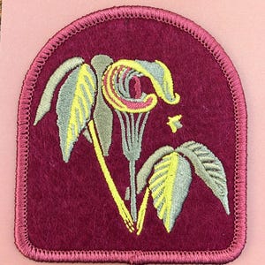 May include: A maroon iron-on patch with an embroidered image of a Jack-in-the-Pulpit flower. The patch has a white border and the text "JACK-IN-THE-PULPIT Spring Wildflower Iron-On Patch" is printed below the image.