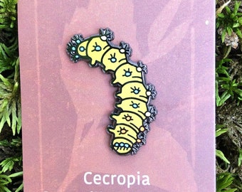 Puede incluir: Un pin de esmalte de oruga Cecropia amarillo y negro en una tarjeta marrón con el texto "Cecropia Caterpillar Enamel Pin" y "SOLSTICEHANDMADE.COM".
