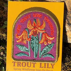 May include: A purple iron-on patch with a gold border featuring three orange and red trout lily flowers with green leaves. The text "Here Today Gone Tomorrow" is embroidered in gold around the flowers. The text "Trout Lily Spring Wildflower Iron-On Patch" is printed below the patch.