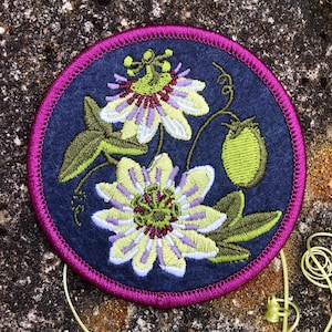 May include: A circular embroidered patch featuring a passion flower design. The patch has a navy blue background with a magenta border. The flower is depicted in shades of yellow, green, and purple, with green leaves and a green fruit. The patch is likely for clothing or accessories.