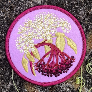 May include: A round embroidered patch featuring a botanical design. The patch has a light purple center with a magenta border. The design includes white flowers, gold leaves, and dark red berries. The patch is set against a textured, dark background.
