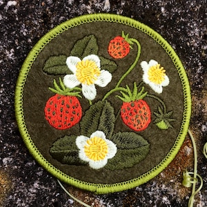 May include: A round, embroidered patch featuring a strawberry design. The patch has a dark green background with a light green border. The design includes red strawberries, white flowers with yellow centers, and green leaves.