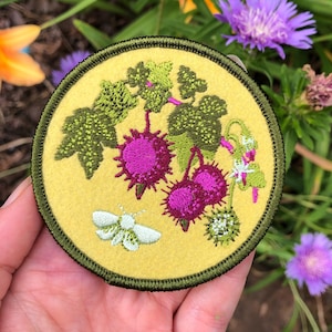 May include: A circular embroidered patch with a yellow background and a dark green border. The design features green leaves, three spiky magenta fruit clusters, and a small pale green moth. The embroidery is detailed with varied stitch textures.