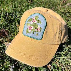 May include: Mustard yellow baseball cap with a floral patch. The patch features white and pink flowers with green leaves on a light blue background. A small bee is also on the patch. The cap is sitting on green grass.