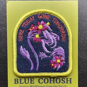 May include: A blue iron-on patch featuring a purple and pink flower design. The patch has the text "Here Today Gone Tomorrow" around the flower design and the text "Blue Cohosh Spring Wildflower Iron-On Patch" below the design.