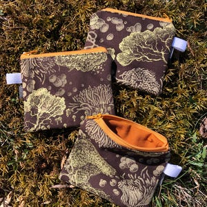 May include: Three brown fabric zipper pouches with a yellow floral pattern and orange zipper pulls. The pouches are lying on a bed of green moss.
