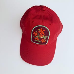 May include: A solid red baseball cap featuring a centered, rounded rectangular patch. The patch has a dark maroon background with an embroidered design of bright orange flowers and a small green hummingbird, outlined in a light mint green border.