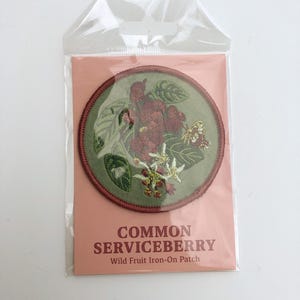 May include: A circular embroidered iron-on patch featuring a botanical illustration of a common serviceberry with green leaves, red berries, white flowers, and a small butterfly, enclosed in a clear plastic sleeve with a peach-colored backing card.