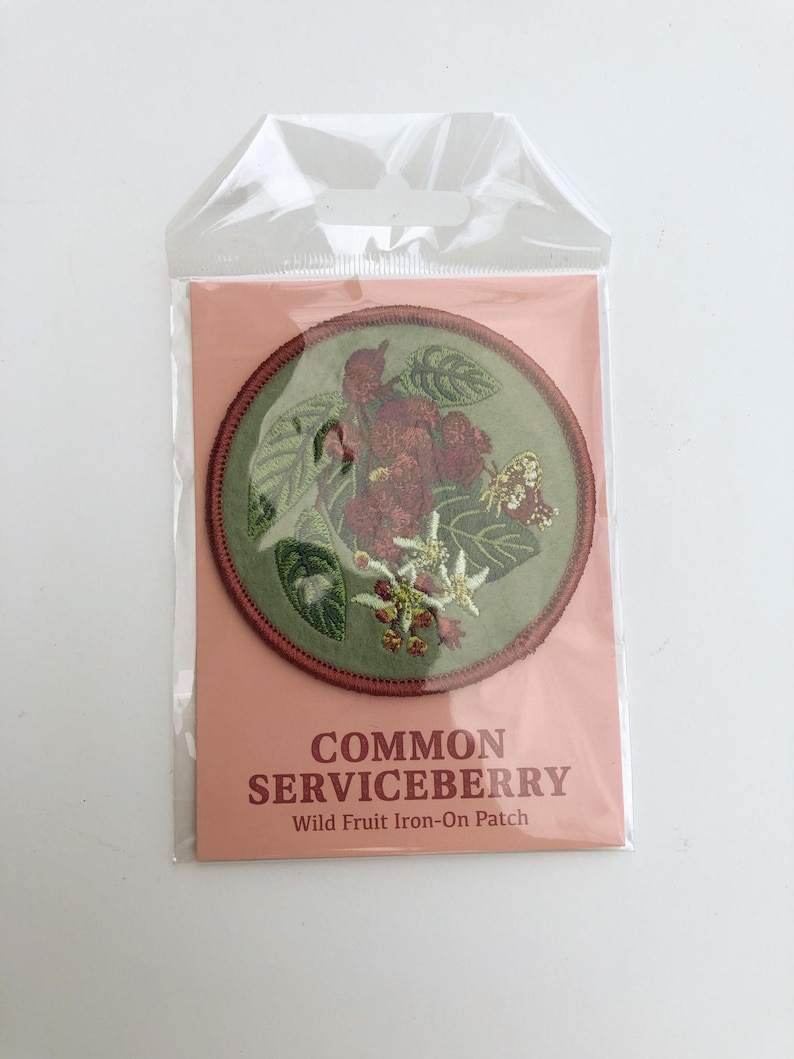 May include: A circular embroidered iron-on patch featuring a botanical illustration of a common serviceberry with green leaves, red berries, white flowers, and a small butterfly, enclosed in a clear plastic sleeve with a peach-colored backing card.