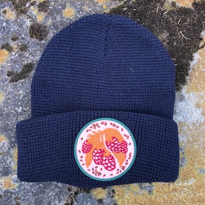May include: A navy blue knitted beanie with a circular embroidered patch featuring a pink and orange stylized image of a berry branch.