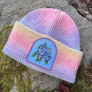 May include: A ribbed knit beanie featuring a pastel gradient of purple, pink, and yellow. A light blue patch is centered on the cuff, displaying an embroidered illustration of blue bell flowers and the text "HERE TODAY GONE TOMORROW" in purple.