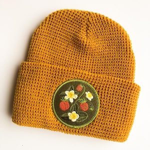 Könnte beinhalten: Eine senfgelbe Strickmütze mit einem runden, aufgestickten Aufnäher auf der Vorderseite. Der Aufnäher zeigt einen dunkelgrünen Hintergrund mit detaillierten Stickereien von roten Erdbeeren, weißen Blüten mit gelber Mitte und grünen Blättern.