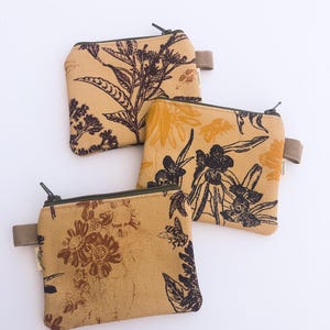 Könnte beinhalten: Drei kleine Reißverschlusstaschen mit botanischen und Insektenmotiven. Die Taschen sind beige mit dunkelbraunen und gelben Blumen- und Bienenmustern. Jede Tasche hat eine kleine Stoffschlaufe und einen dunklen Reißverschluss.