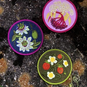May include: Three embroidered patches with floral designs. One features passion flowers on a navy background, another showcases elderberries on a pink background, and the third displays strawberries on a green background. Each patch is round with a contrasting border.