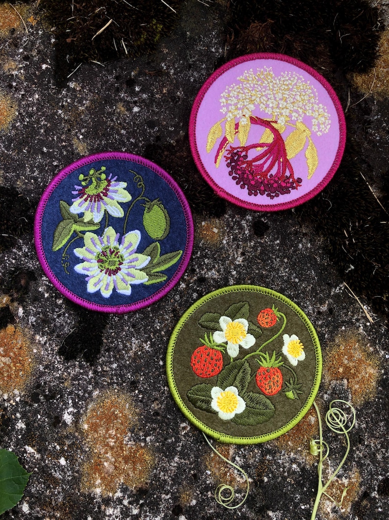 May include: Three embroidered patches with floral designs. One features passion flowers on a navy background, another showcases elderberries on a pink background, and the third displays strawberries on a green background. Each patch is round with a contrasting border.