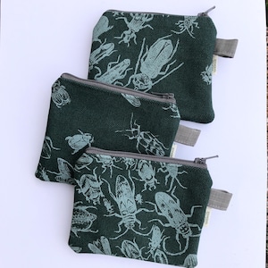 May include: Three dark green zippered pouches with a light blue insect pattern. Each pouch has a grey zip and a small grey fabric loop. The pouches are arranged in a slightly overlapping pattern.
