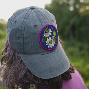 May include: A charcoal gray baseball cap featuring a circular embroidered patch on the front. The patch has a dark blue background with a vibrant purple border and displays detailed floral imagery of passion flowers in shades of white and purple.