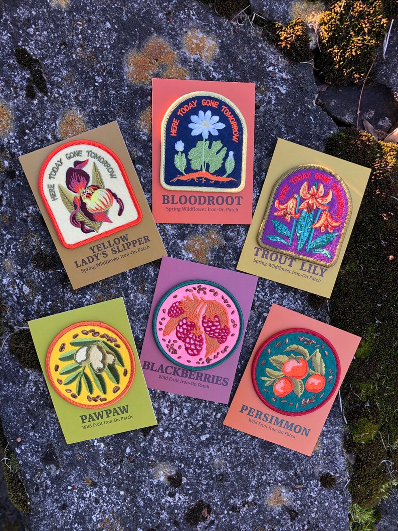 K&ouml;nnte beinhalten: Zehn gestickte B&uuml;gelbilder mit verschiedenen Fr&uuml;hlingswildblumen und Wildfr&uuml;chten. Die Patches sind rund und haben eine farbenfrohe, Vintage-&Auml;sthetik. Der Text auf den Patches lautet: "Here Today Gone Tomorrow", "Yellow Lady's Slipper", "Bloodroot", "Trout Lily", "Pawpaw", "Blackberries", "Persimmon".