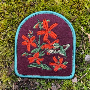 May include: Embroidered patch featuring a floral design with a hummingbird. The patch has a burgundy background with orange flowers and green leaves. The patch is outlined with a teal border.