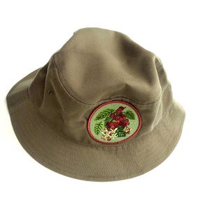 May include: A solid olive-green bucket hat featuring a circular embroidered patch on the front. The patch displays a detailed illustration of red coffee cherries, green leaves, and white coffee blossoms, all bordered by a thin red stitched edge.